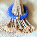 A beaded bracelet in bright cobalt blue with a single gold accent bead, displayed on a textured beige jute cone stand against a soft beige fabric background.