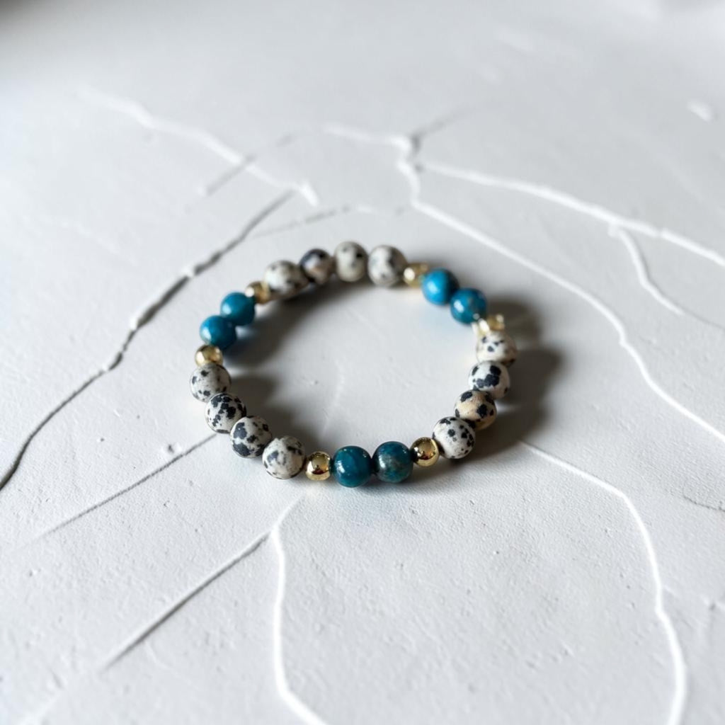 A beaded stretch bracelet featuring white and black speckled beads, bright blue beads, and small gold spacer beads, displayed on a textured white surface.