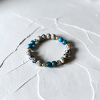 A beaded stretch bracelet featuring white and black speckled beads, bright blue beads, and small gold spacer beads, displayed on a textured white surface.