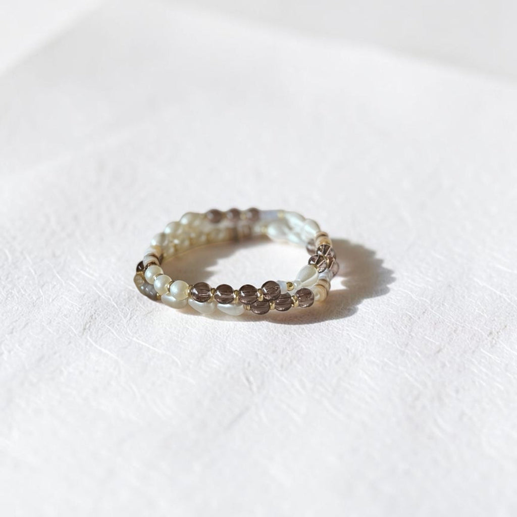 A beaded stretch bracelet featuring small white pearls and brown smoky crystal beads, resting on a textured white surface with a soft shadow.