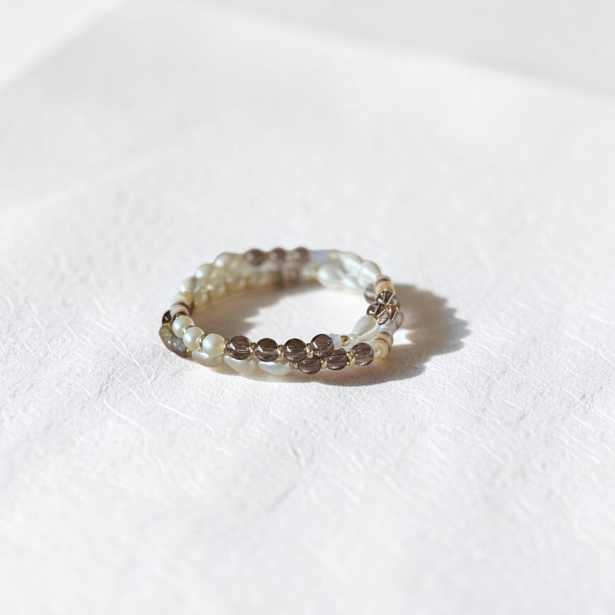 A beaded stretch bracelet featuring small white pearls and brown smoky crystal beads, resting on a textured white surface with a soft shadow.