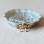 A delicate gold wire bow earring with clear faceted beads, displayed inside a small, scalloped light gray ceramic dish on a textured beige surface.