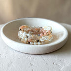 A stack of beaded stretch bracelets with white baroque pearls, peach-pink beads, and small metallic accents, displayed on a small round ceramic dish.