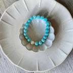 A turquoise beaded bracelet with a small white pearl accent, displayed on a small scalloped cream dish over a larger matching plate on a light wood surface.