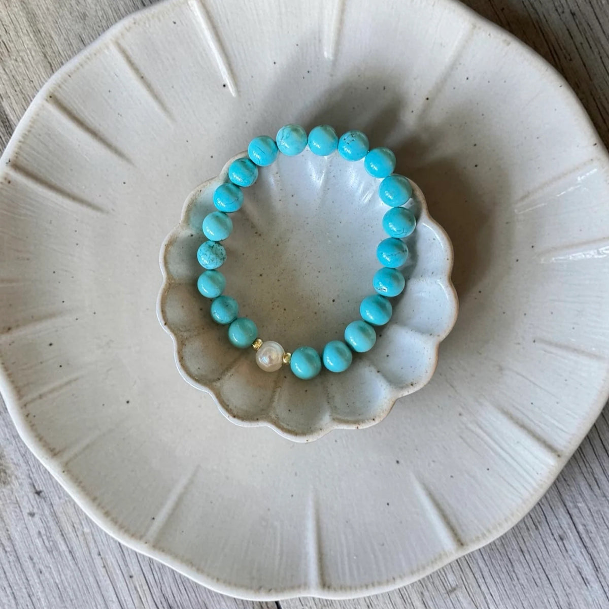 A turquoise beaded bracelet with a small white pearl accent, displayed on a small scalloped cream dish over a larger matching plate on a light wood surface.