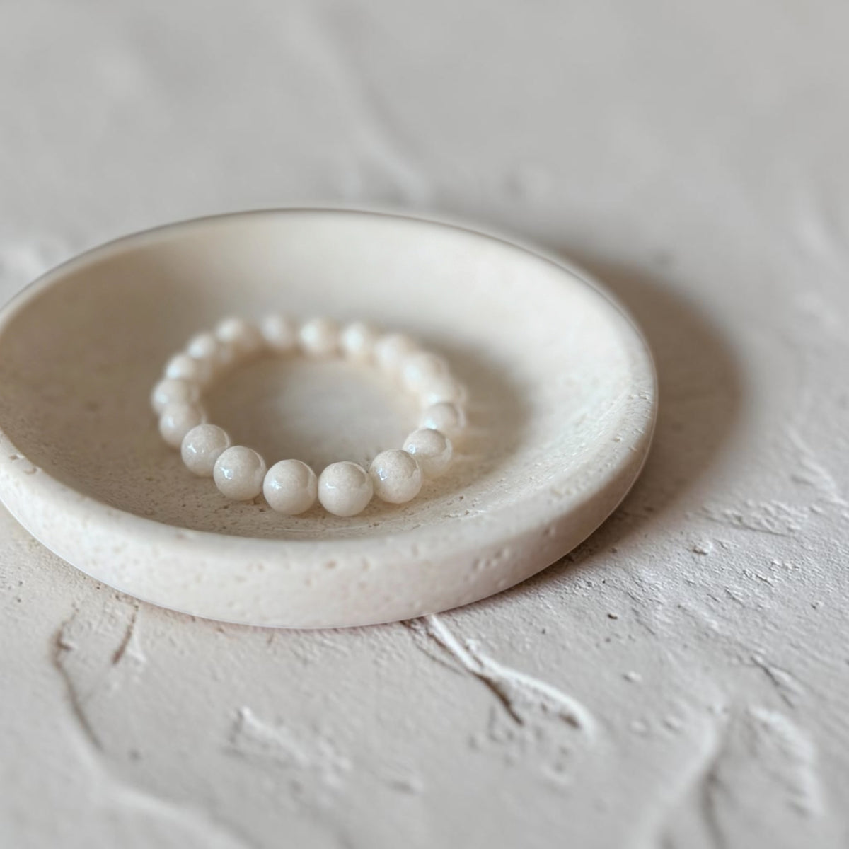 A beaded stretch bracelet made of 10mm cream beads, displayed inside a shallow round ceramic dish on a textured light gray surface.