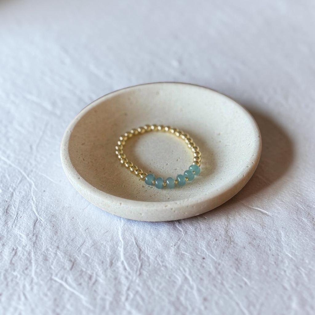A delicate gold beaded ring with small light blue gemstone accents, displayed on a small round ceramic dish over a textured white fabric background.
