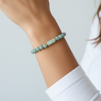 A close-up of a person's wrist wearing a beaded bracelet made of pale mint green beads with one larger central bead, against a soft light gray background.