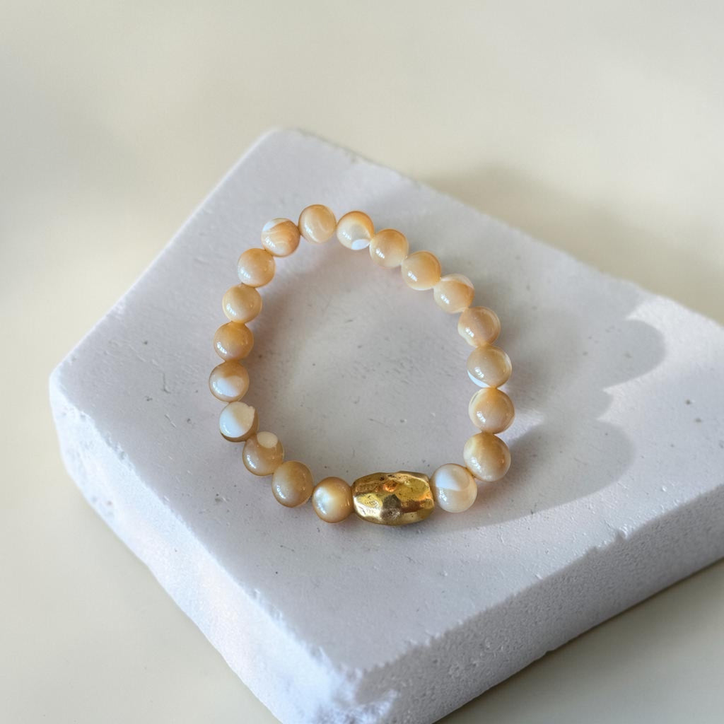 A beaded bracelet made of warm cream and white mother of pearls with a faceted gold accent accent bead, displayed on a white textured block.