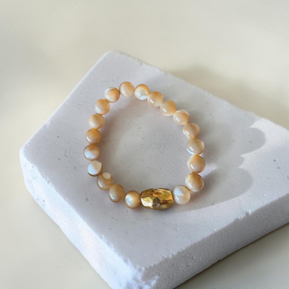A beaded bracelet made of warm cream and white mother of pearls with a faceted gold accent accent bead, displayed on a white textured block.