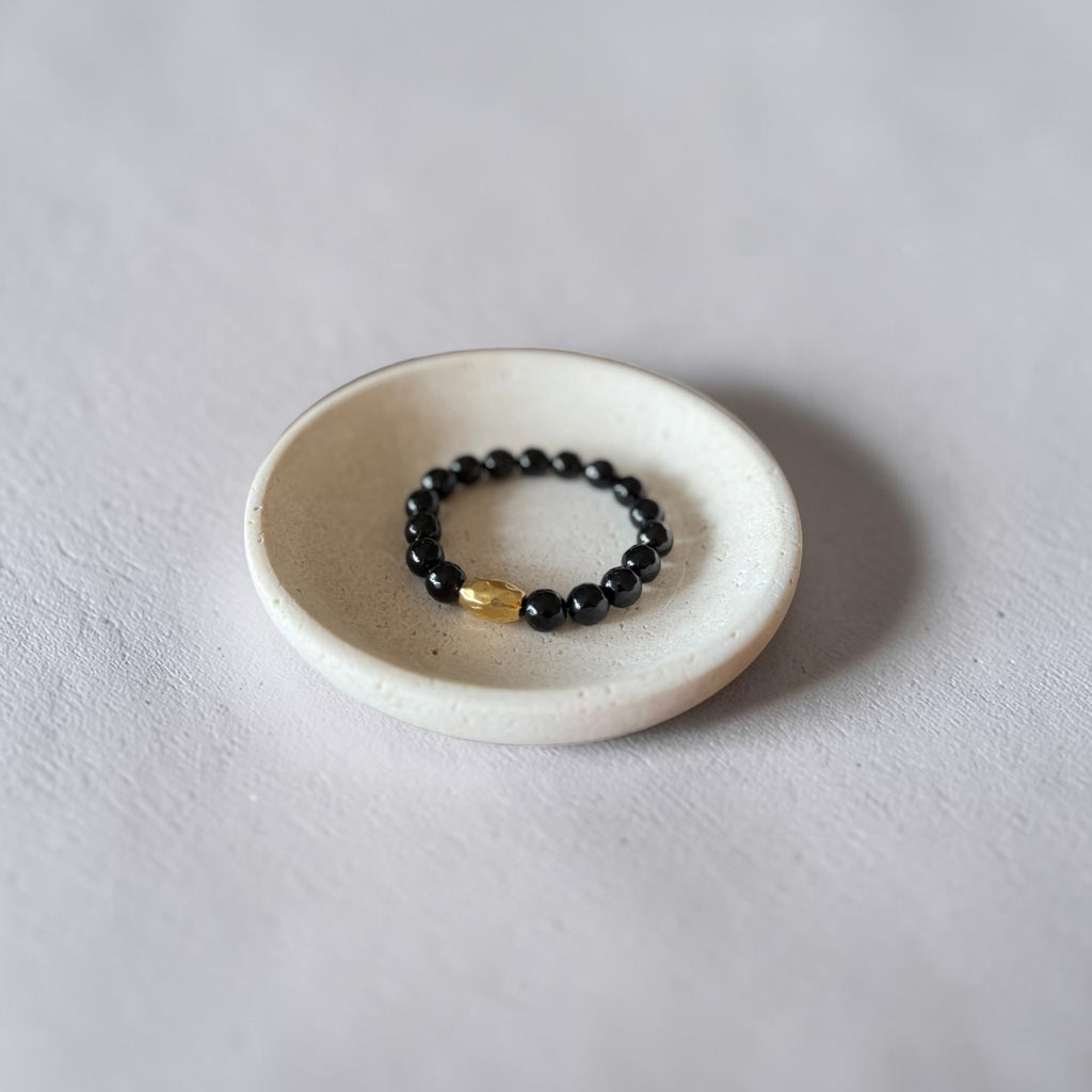 A beaded stretch bracelet with glossy black beads and a single gold accent bead, displayed inside a small white ceramic dish on a light gray textured surface.