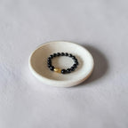 A beaded stretch bracelet with glossy black beads and a single gold accent bead, displayed inside a small white ceramic dish on a light gray textured surface.