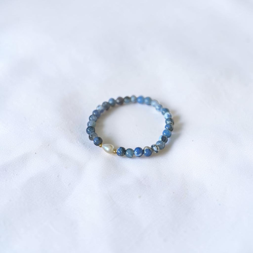 A beaded stretch bracelet featuring round blue and gray beads with a single white pearl accent, resting on a white fabric surface.