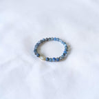 A beaded stretch bracelet featuring round blue and gray beads with a single white pearl accent, resting on a white fabric surface.