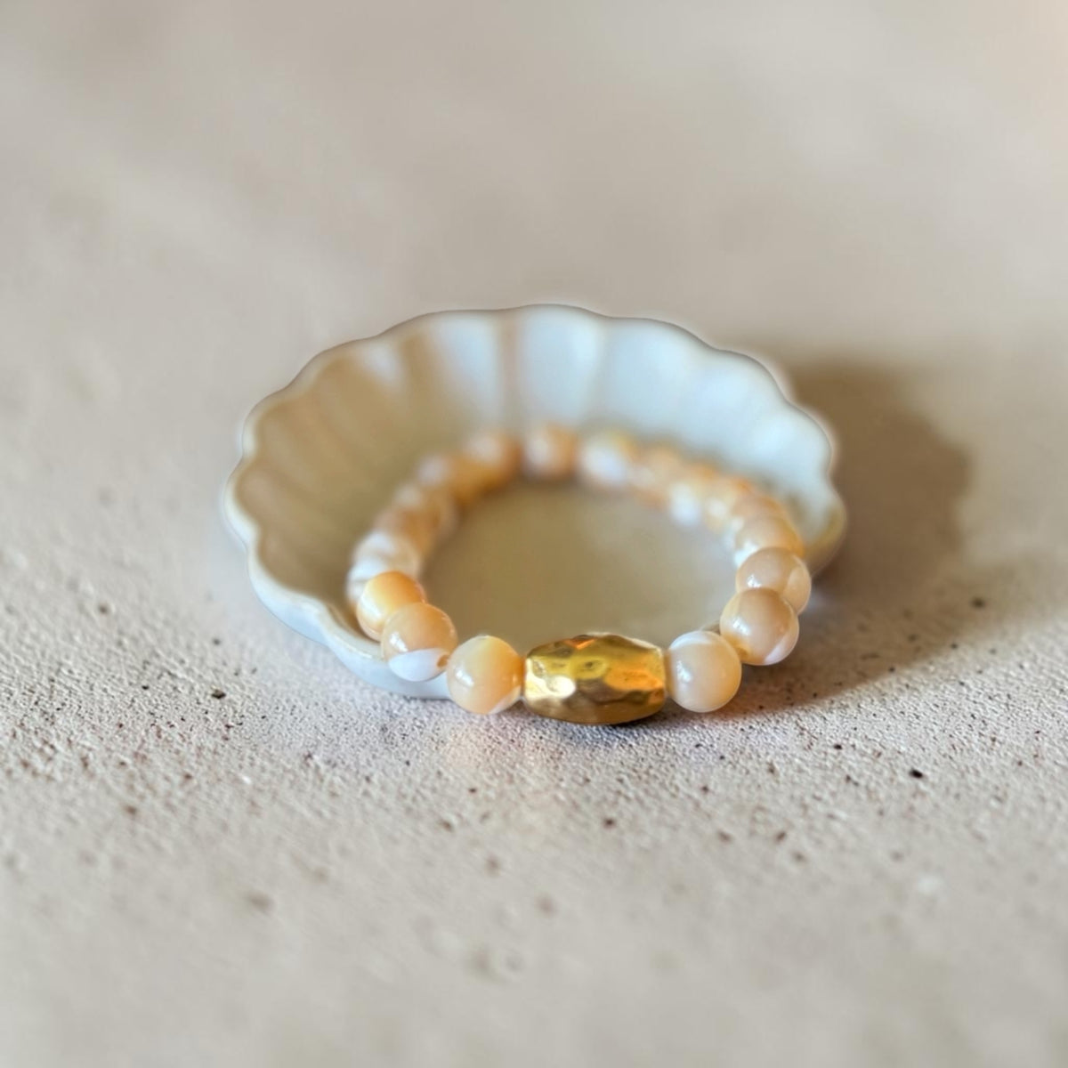 A beaded stretch bracelet with white pearl-like round beads and a central faceted gold oval bead, displayed on a small white scalloped dish over a textured beige surface.