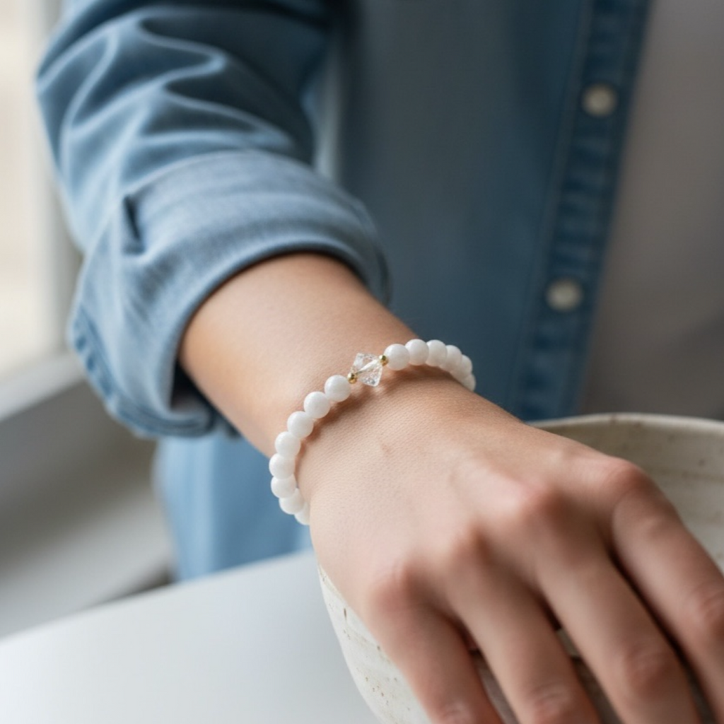 A close-up of a person's wrist wearing a white beaded bracelet with a small gold-toned charm, paired with a light blue denim shirt.