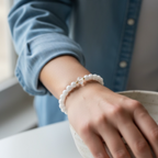 A close-up of a person's wrist wearing a white beaded bracelet with a small gold-toned charm, paired with a light blue denim shirt.