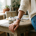 A person in a cream linen shirt and blue jeans rests their hand on a rustic wooden table in a sunlit studio, wearing a beaded bracelet with a blue and white patterned center.