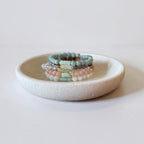 A stack of beaded bracelets in pastel mint green, peach, white, and brown tones displayed on a small round ceramic dish against a light neutral background.