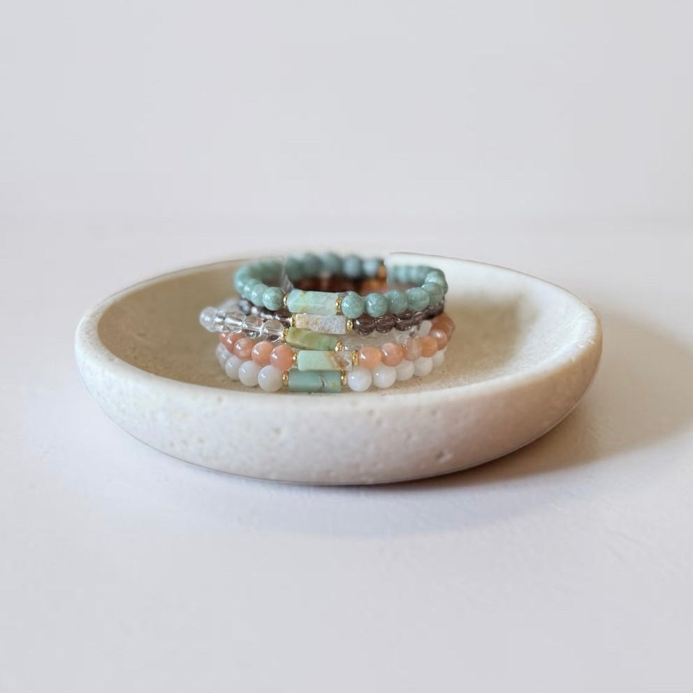 A stack of beaded bracelets in pastel mint green, peach, white, and brown tones displayed on a small round ceramic dish against a light neutral background.