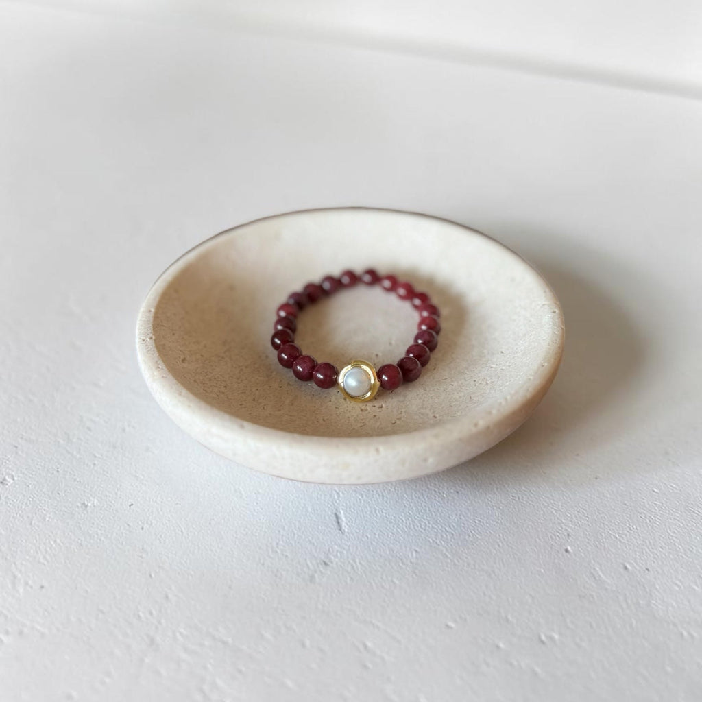 8mm Red Garnet Jade Bead Bracelet with a Gold And Pearl Accent. Accent has a round pearl with gold surrounding the pearl itself. This bracelet is sitting in a dish.
