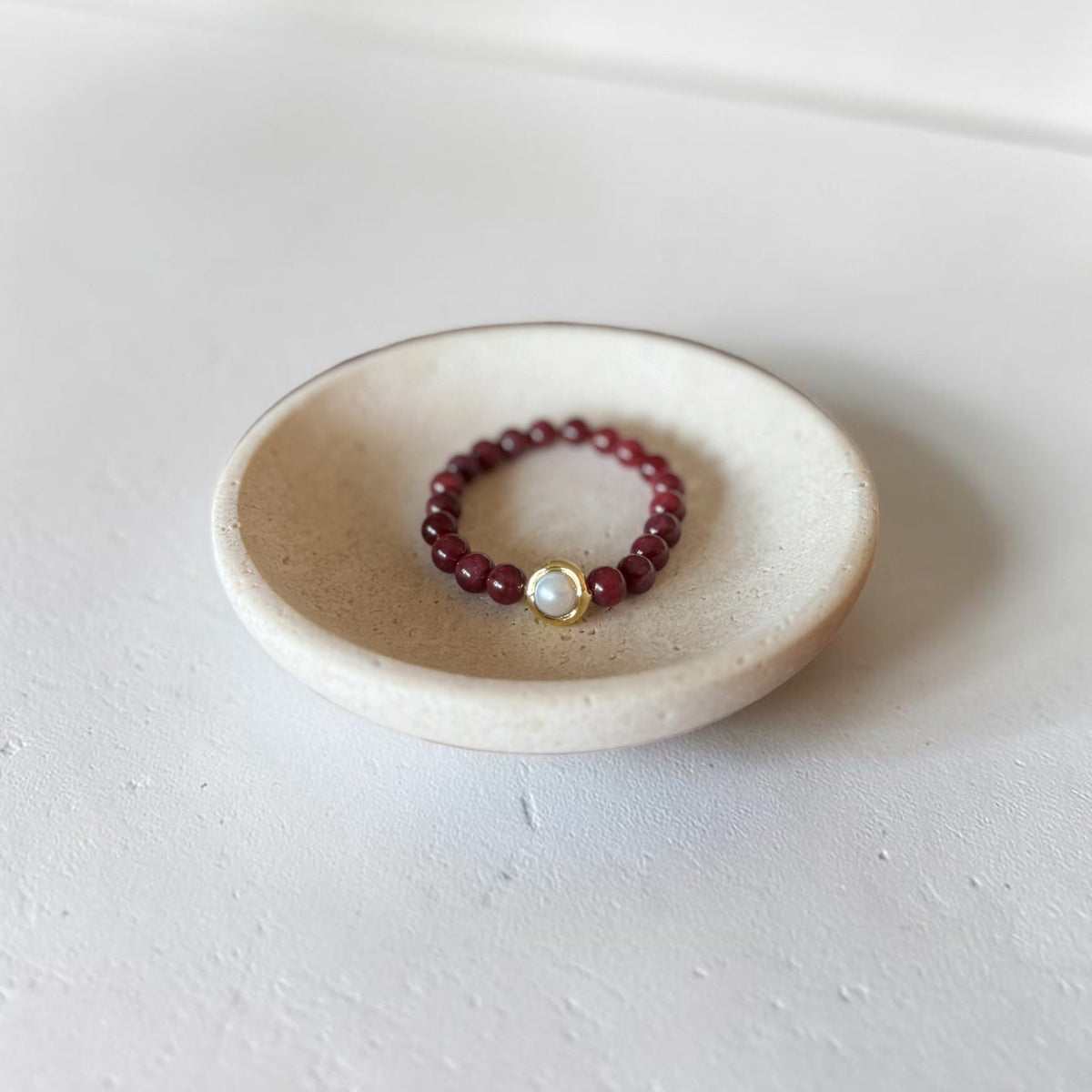 8mm Red Garnet Jade Bead Bracelet with a Gold And Pearl Accent. Accent has a round pearl with gold surrounding the pearl itself. This bracelet is sitting in a dish.