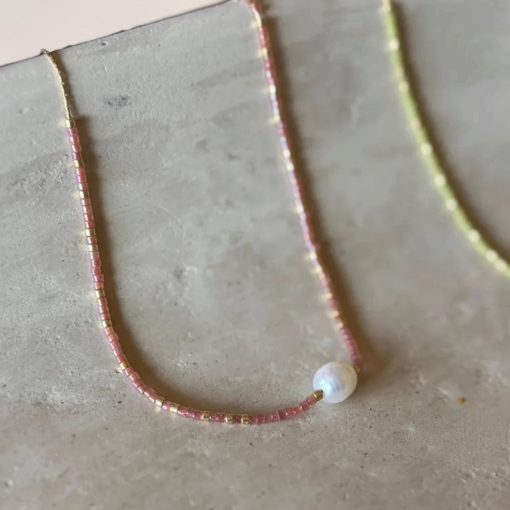 A delicate beaded necklace with small pink and gold beads and a single white pearl bead, displayed on a textured gray surface.