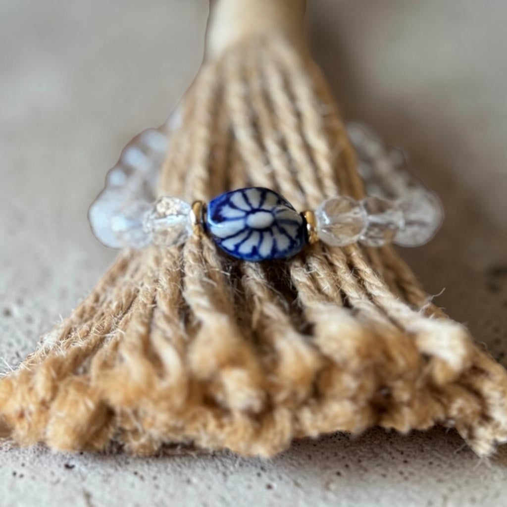 A handmade beaded ring featuring a central blue and white floral bead, flanked by clear faceted glass beads, resting on a rustic jute rope prop.