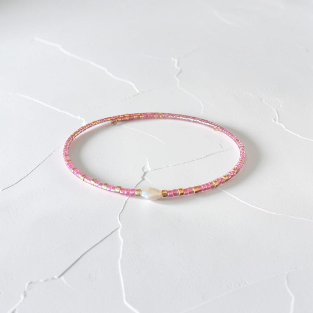 A delicate pink beaded bracelet with small gold and white accents, resting on a white textured surface.