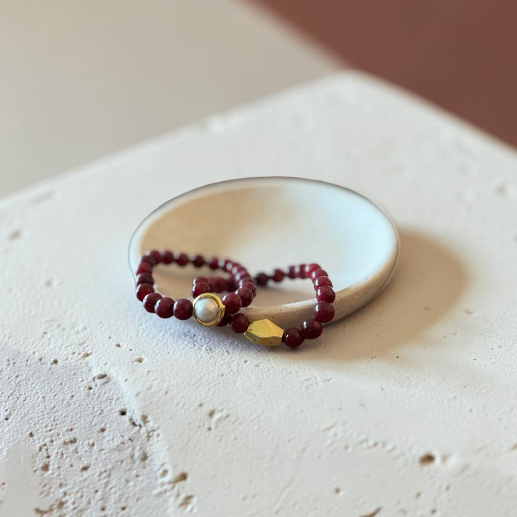 A beaded bracelet with deep red beads, a white pearl, and a gold geometric accent, displayed on a small white dish over a textured beige surface.