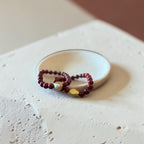 A beaded bracelet with deep red beads, a white pearl, and a gold geometric accent, displayed on a small white dish over a textured beige surface.