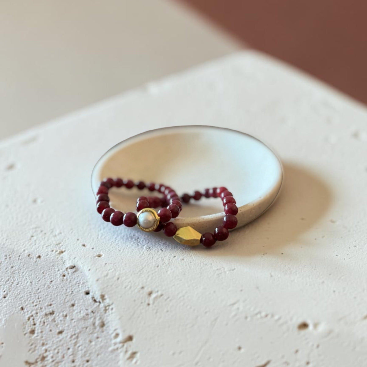 A beaded bracelet with deep red beads, a white pearl, and a gold geometric accent, displayed on a small white dish over a textured beige surface.