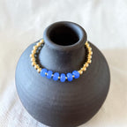 A beaded bracelet with bright blue oval beads and small gold spacer beads, displayed around a matte black ceramic vase on a light fabric surface.
