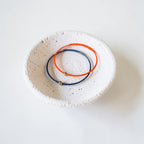 Two thin wire bracelets, one orange and one navy blue, each with a small metallic bead, displayed inside a small speckled white ceramic dish on a white background.