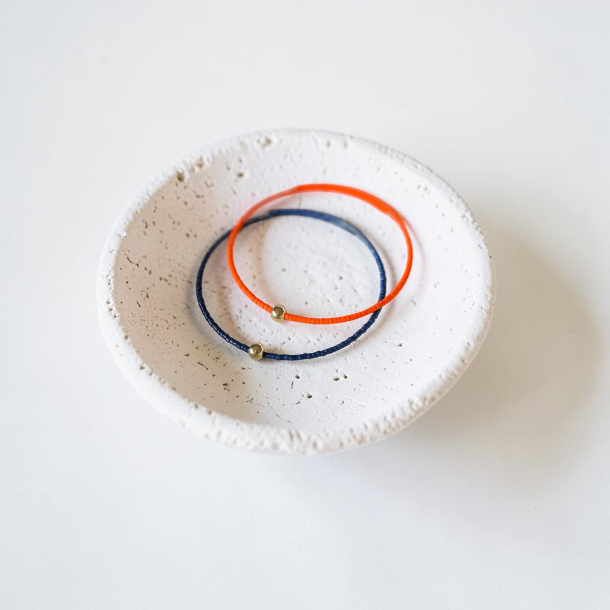 Two thin wire bracelets, one orange and one navy blue, each with a small metallic bead, displayed inside a small speckled white ceramic dish on a white background.