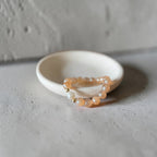A beaded stretch bracelet with peach-orange and white beads, resting on a small white ceramic dish against a textured gray concrete background.