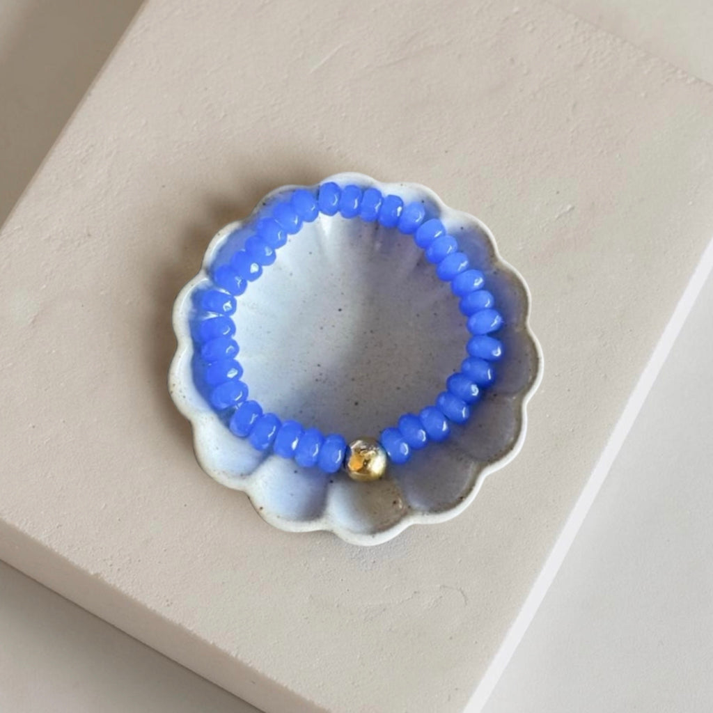 A beaded bracelet with small blue beads and a single gold accent bead, displayed on a scalloped white dish over a beige surface.