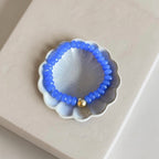A beaded bracelet with small blue beads and a single gold accent bead, displayed on a scalloped white dish over a beige surface.