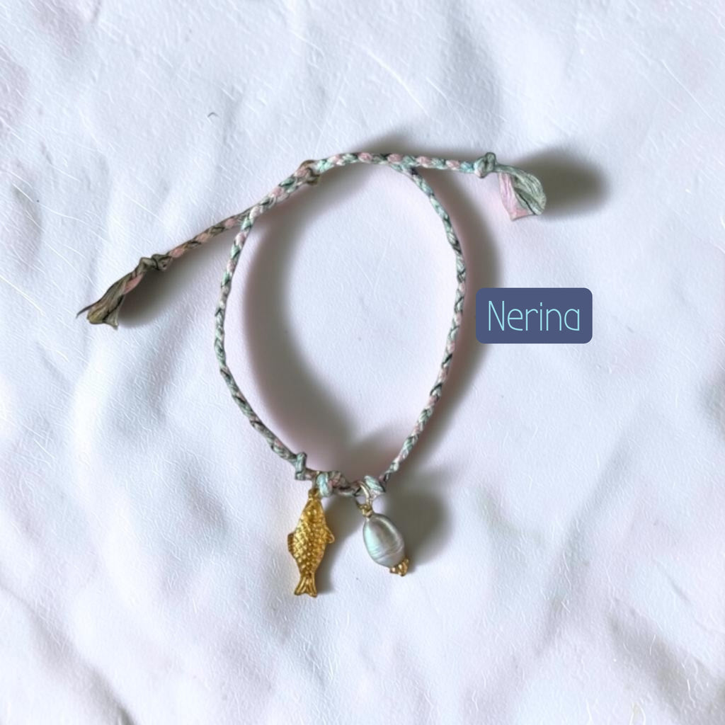 A delicate braided string bracelet with a small gold fish charm and a white shell charm, laid flat on a white fabric background. A blue label with the text 'Nerina' is placed next to the bracelet.