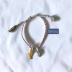 A delicate braided string bracelet with a small gold fish charm and a white shell charm, laid flat on a white fabric background. A blue label with the text 'Nerina' is placed next to the bracelet.