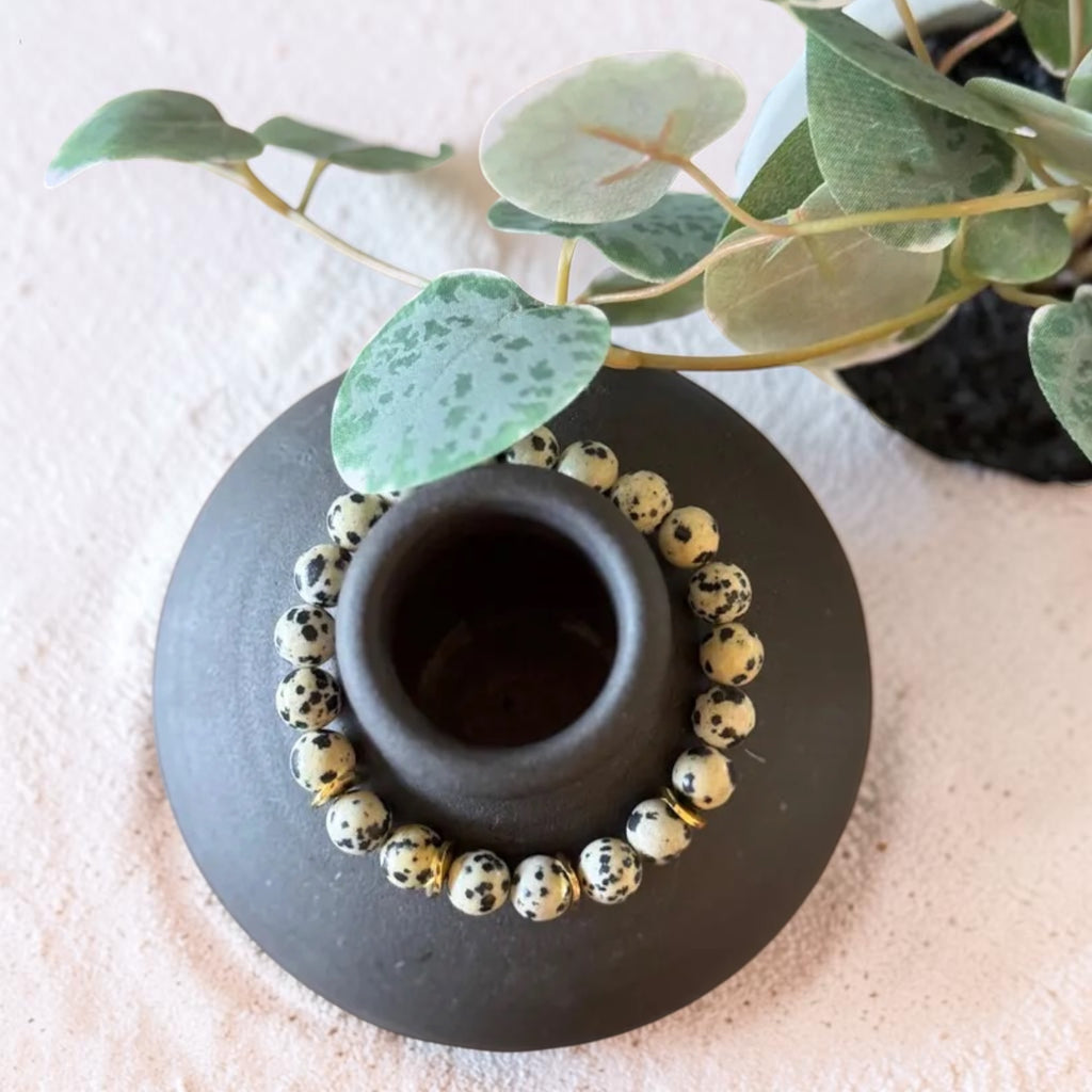 A beaded bracelet with speckled cream and black beads arranged around the rim of a small matte black ceramic cup, styled with green leaves on a light textured surface.