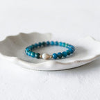 A beaded stretch bracelet with turquoise-blue and teal beads, a small white pearl, and a tiny gold spacer, displayed on a white ceramic dish against a light gray background.