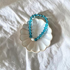A beaded stretch bracelet made of faceted turquoise-blue beads with a small white bead accent, displayed on a small white scalloped dish over a white fabric background.