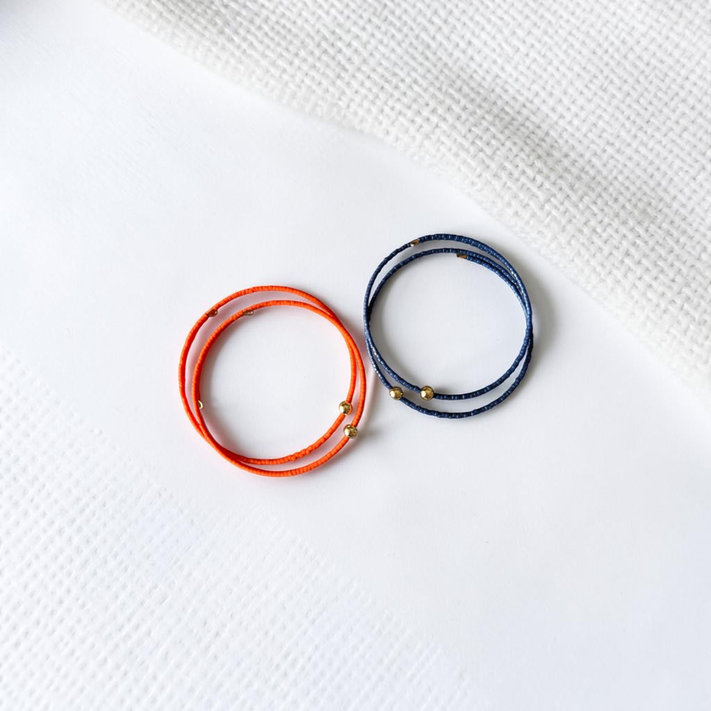 Two thin wrap-style bracelets, one bright orange and one dark blue, each with small gold bead accents, arranged on a white textured surface.