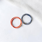 Two thin wrap-style bracelets, one bright orange and one dark blue, each with small gold bead accents, arranged on a white textured surface.
