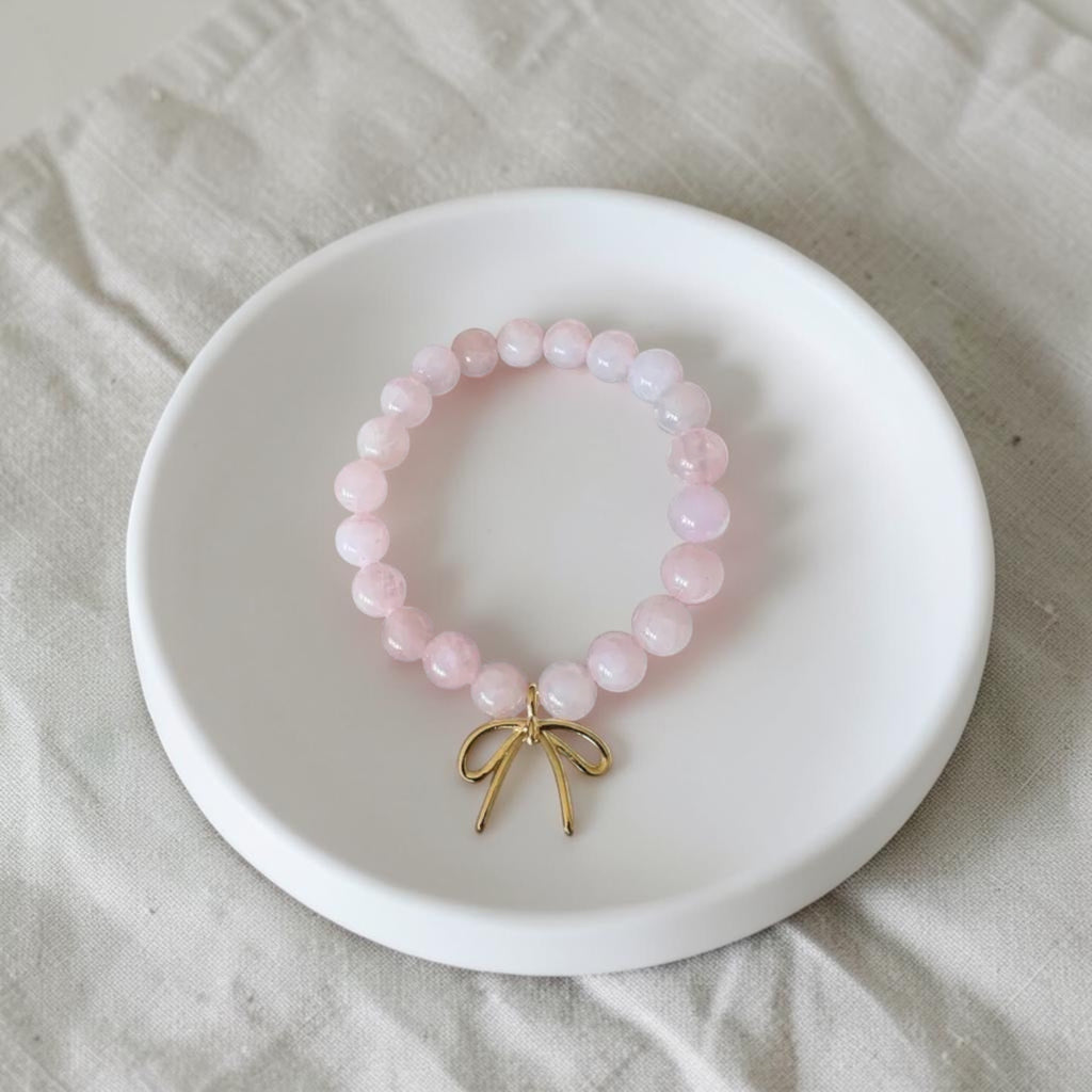 A pink beaded bracelet with a small gold bow charm, displayed on a white ceramic dish over a light beige fabric background.
