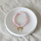 A pink beaded bracelet with a small gold bow charm, displayed on a white ceramic dish over a light beige fabric background.
