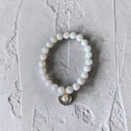 A beaded stretch bracelet featuring white pearl-like round beads and a small gold-toned shell charm with a pearl center, displayed on a textured light gray surface.