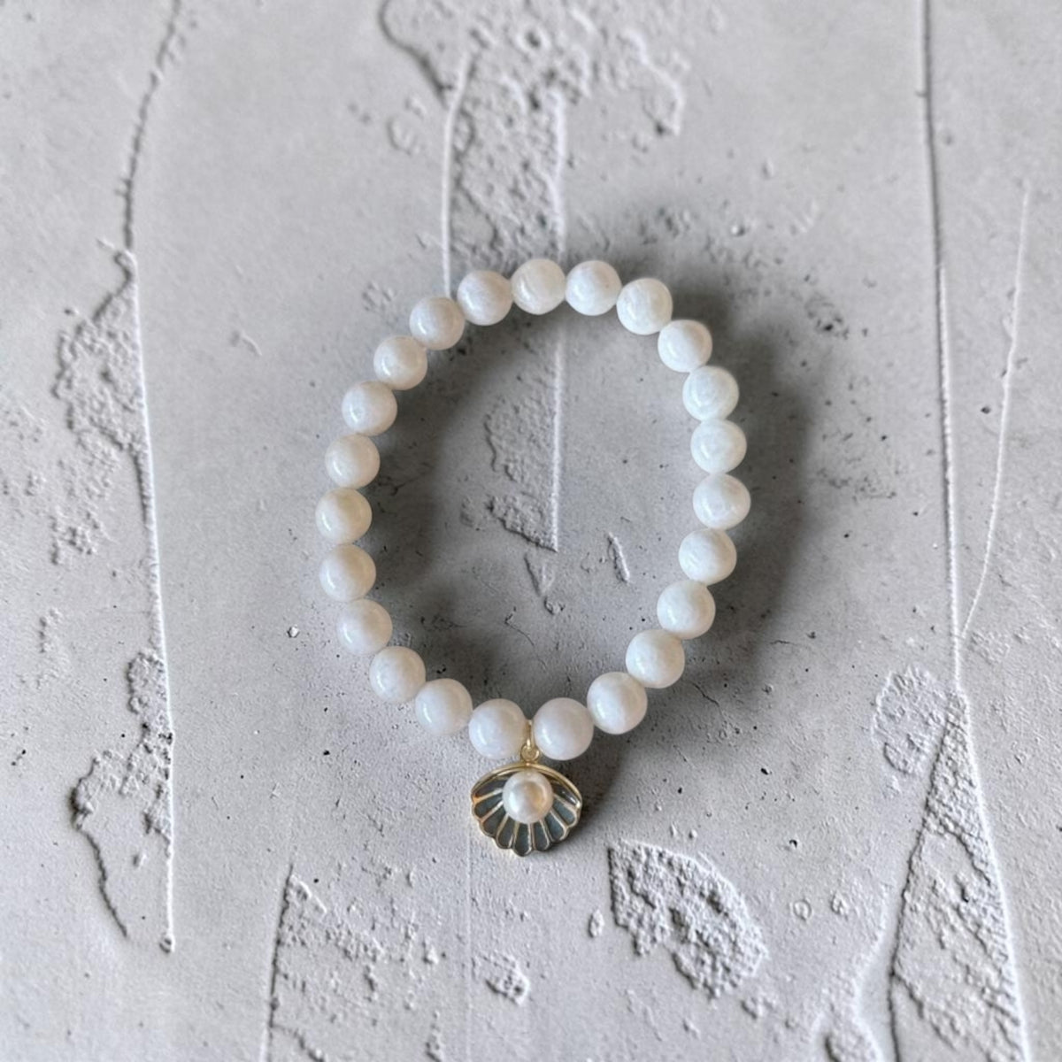 A beaded stretch bracelet featuring white pearl-like round beads and a small gold-toned shell charm with a pearl center, displayed on a textured light gray surface.