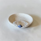 A beaded stretch bracelet with clear faceted beads and a central blue and white patterned shell-shaped bead, displayed in a small textured ceramic dish on a light gray surface.